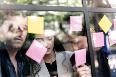 İş adamları ofisteki cam pencere duvarına yapışkan notlar yapıştırarak beyin fırtınası yapıyorlar. Pencere camı gün ışığını ve çevreyi yansıtıyor. Yeni fikir için yaratıcı grup girişimi iş toplantısı.