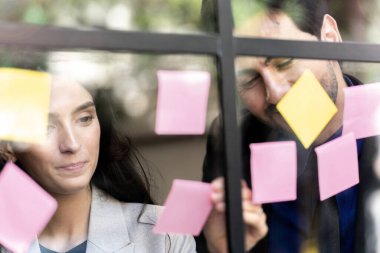 İş adamları ofisteki cam pencere duvarına yapışkan notlar yapıştırarak beyin fırtınası yapıyorlar. Pencere camı gün ışığını ve çevreyi yansıtıyor. Yeni fikir için yaratıcı grup girişimi iş toplantısı.