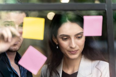 İş kadını düşünce ve beyin fırtınası ofisindeki cam pencere duvarına yapışkan notlar yapıştırıyor. Pencere camı gün ışığını ve doğayı yansıtıyor. Yeni fikir için yaratıcı grup girişimi iş toplantısı.