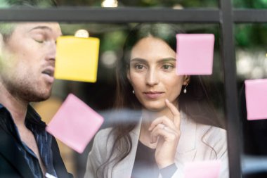 İş kadını düşünce ve beyin fırtınası ofisindeki cam pencere duvarına yapışkan notlar yapıştırıyor. Pencere camı gün ışığını ve doğayı yansıtıyor. Yeni fikir için yaratıcı grup girişimi iş toplantısı.