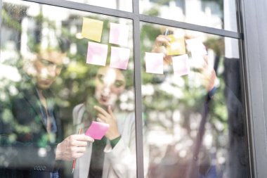 İş adamları ofisteki cam pencere duvarına yapışkan notlar yapıştırarak beyin fırtınası yapıyorlar. Pencere camı gün ışığını ve çevreyi yansıtıyor. Yeni fikir için yaratıcı grup girişimi iş toplantısı.
