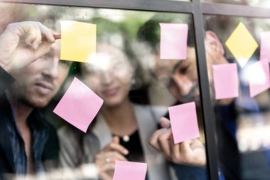 İş adamları ofisteki cam pencere duvarına yapışkan notlar yapıştırarak beyin fırtınası yapıyorlar. Pencere camı gün ışığını ve çevreyi yansıtıyor. Yeni fikir için yaratıcı grup girişimi iş toplantısı.