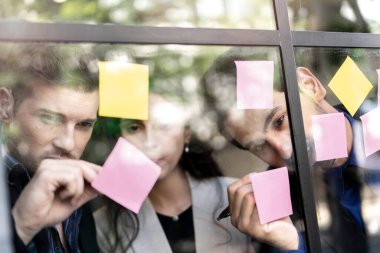 İş adamları ofisteki cam pencere duvarına yapışkan notlar yapıştırarak beyin fırtınası yapıyorlar. Pencere camı gün ışığını ve çevreyi yansıtıyor. Yeni fikir için yaratıcı grup girişimi iş toplantısı.