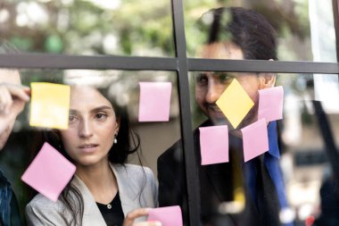 İş adamları ofisteki cam pencere duvarına yapışkan notlar yapıştırarak beyin fırtınası yapıyorlar. Pencere camı gün ışığını ve çevreyi yansıtıyor. Yeni fikir için yaratıcı grup girişimi iş toplantısı.