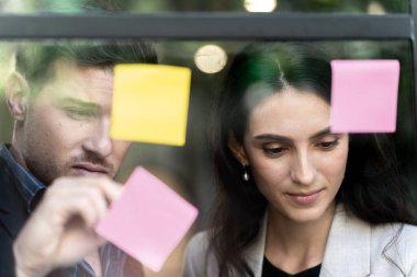 İş kadını düşünce ve beyin fırtınası ofisindeki cam pencere duvarına yapışkan notlar yapıştırıyor. Pencere camı gün ışığını ve doğayı yansıtıyor. Yeni fikir için yaratıcı grup girişimi iş toplantısı.