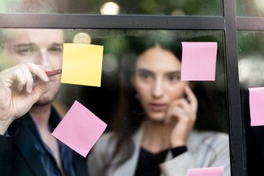 İş kadını düşünce ve beyin fırtınası ofisindeki cam pencere duvarına yapışkan notlar yapıştırıyor. Pencere camı gün ışığını ve doğayı yansıtıyor. Yeni fikir için yaratıcı grup girişimi iş toplantısı.