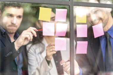 İş adamları ofisteki cam pencere duvarına yapışkan notlar yapıştırarak beyin fırtınası yapıyorlar. Pencere camı gün ışığını ve çevreyi yansıtıyor. Yeni fikir için yaratıcı grup girişimi iş toplantısı.