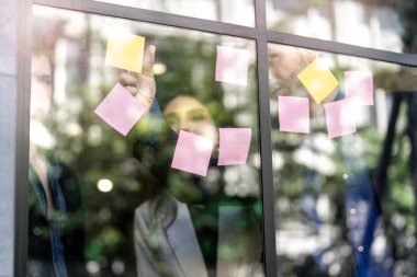 İş adamları ofisteki cam pencere duvarına yapışkan notlar yapıştırarak beyin fırtınası yapıyorlar. Pencere camı gün ışığını ve çevreyi yansıtıyor. Yeni fikir için yaratıcı grup girişimi iş toplantısı.