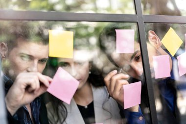 İş adamları ofisteki cam pencere duvarına yapışkan notlar yapıştırarak beyin fırtınası yapıyorlar. Pencere camı gün ışığını ve çevreyi yansıtıyor. Yeni fikir için yaratıcı grup girişimi iş toplantısı.