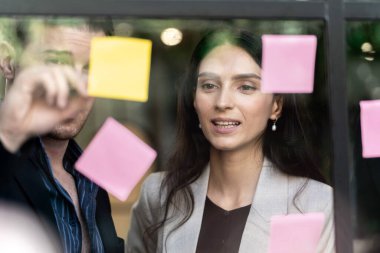 İş kadını düşünce ve beyin fırtınası ofisindeki cam pencere duvarına yapışkan notlar yapıştırıyor. Pencere camı gün ışığını ve doğayı yansıtıyor. Yeni fikir için yaratıcı grup girişimi iş toplantısı.