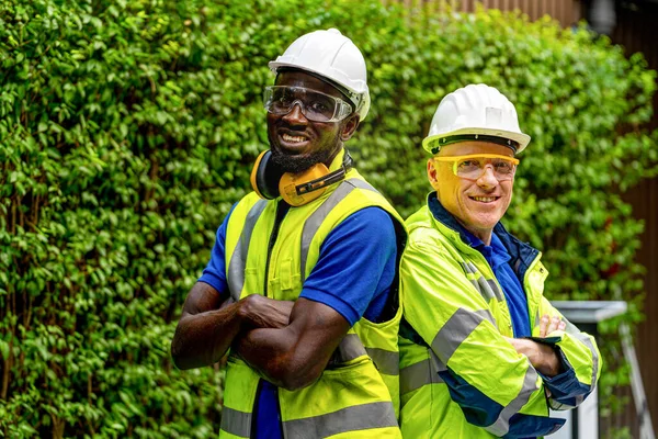 team factory worker technician engineer men standing confidence with green working suite dress and safety helmet. Background of green trees wall.