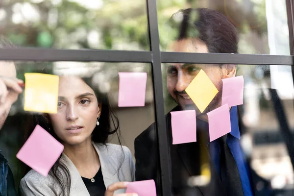 İş adamları ofisteki cam pencere duvarına yapışkan notlar yapıştırarak beyin fırtınası yapıyorlar. Pencere camı gün ışığını ve çevreyi yansıtıyor. Yeni fikir için yaratıcı grup girişimi iş toplantısı.