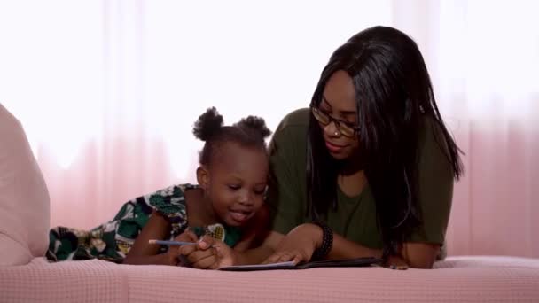 幸せな家族アフリカの母親と子供が一緒に勉強しながら ベッドルームでベッドに横になります アフリカの少女が絵を描くとき 母親は彼女に教える — ストック動画