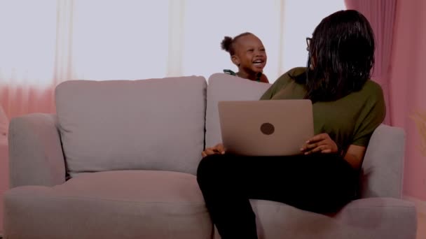 アフリカの母親はラップトップコンピュータで作業し ソファの椅子に座っています 女の子はお母さんの周りを歩き お母さんと遊んでいる間 家庭で働くこと 家族を大切にすることをコンセプトに — ストック動画