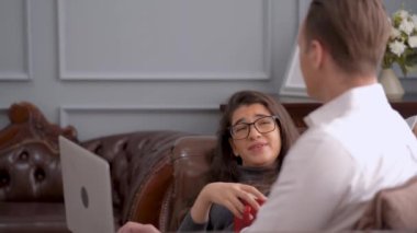 Rahatlatıcı kadın kanepeye uzanıp erkeklerle konuşur. Adam dizüstü bilgisayar kullanıyor ve genç bir kadının yanına oturuyor.