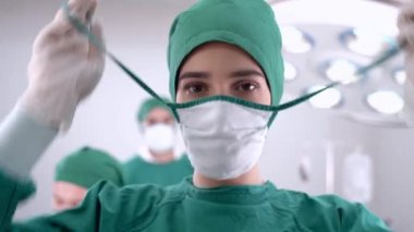 Cerrah kadın doktor gülümseyip tıbbi yüz korumasını kaldırırken, personel ve doktor da tıbbi tesiste çalışıyor. Ameliyat odasındaki ışıklandırma ekipmanlarıyla..