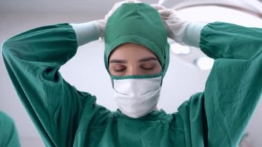 Cerrah kadın doktor gülümseyip tıbbi yüz korumasını kaldırırken, personel ve doktor da tıbbi tesiste çalışıyor. Ameliyat odasındaki ışıklandırma ekipmanlarıyla..