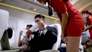 Uçak koltuğunda oturduktan sonra maske takan bir iş adamı. Kadın uçuş görevlisi ya da kabin görevlisi yolcu koltuklarını ve ekipmanları kontrol ediyor..