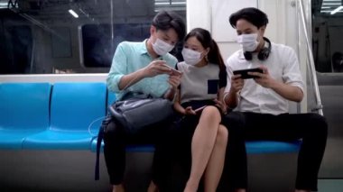 Asyalı erkek ve kadın yüz maskeli metro istasyonunda oturuyor ve toplu taşıma araçlarıyla seyahat eden insanlarla akıllı telefon kullanıyor. Salgın Hastalıklı Corona Virüsü ile yeni bir normal yaşam.