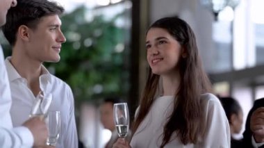 Ortak başarılarını kutluyorlar. Kadın ve erkek neşeli iş adamları şampanya içip sohbet ederken diğer insanlar arka planda iletişim kuruyor..