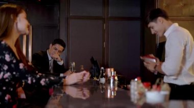 İnsanlar gece kulübünde kutlamak için tezgahta oturup kokteyl içiyorlar. Genç adam, gülümseyen barmen profesyonel olarak şişeyi kullanarak içkiyi karıştırır..