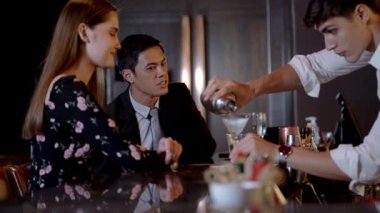 İnsanlar gece kulübünde kutlamak için tezgahta oturup kokteyl içiyorlar. Genç adam, gülümseyen barmen profesyonel olarak şişeyi kullanarak içkiyi karıştırır..