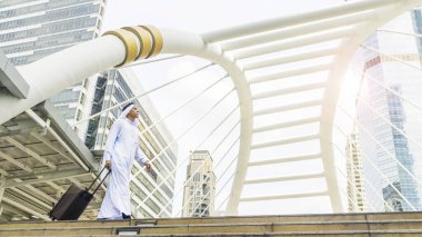 İş adamı Arap gezgin bir bavul taşıyor ve modern çelik yapısı ve dış duvarı modern binasıyla iş yeri simgesi olan şehirde yürüyor. 