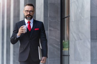 Takım elbiseli üst düzey yönetici elinde bir fincan kahve ve dizüstü bilgisayar tutuyor. Sabahın köründe kahve saatinde iş hayatı. Taze ve mutlu hisseden adam, başarısından emin..