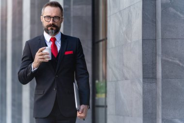 Takım elbiseli üst düzey yönetici elinde bir fincan kahve ve dizüstü bilgisayar tutuyor. Sabahın köründe kahve saatinde iş hayatı. Taze ve mutlu hisseden adam, başarısından emin..