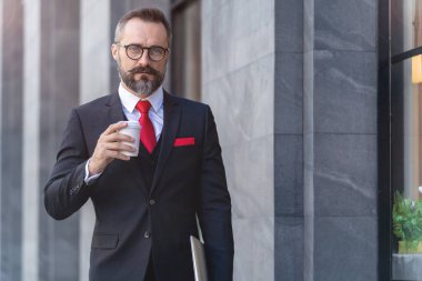 Takım elbiseli üst düzey yönetici elinde bir fincan kahve ve dizüstü bilgisayar tutuyor. Sabahın köründe kahve saatinde iş hayatı. Taze ve mutlu hisseden adam, başarısından emin..