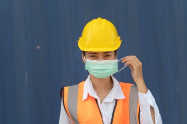 Asyalı kadın işçi mavi duvarda duruyor. İşçi kadın işçi koruyucu maske ve güvenlik kaskı takıyor ve turuncu yansıtıcı güvenlik elbisesi giyiyor. 