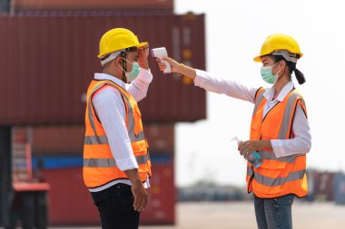 Yüz maskesi ve güvenlik elbisesi takan fabrika kadın çalışanı, temassız kızılötesi termometreyle kuyrukta bekleyen işçi sıcaklığını ölçüyor. kargo konteynırının arkaplanı.