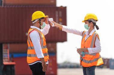 Yüz maskesi ve güvenlik elbisesi takan fabrika kadın çalışanı, temassız kızılötesi termometreyle kuyrukta bekleyen işçi sıcaklığını ölçüyor. kargo konteynırının arkaplanı.