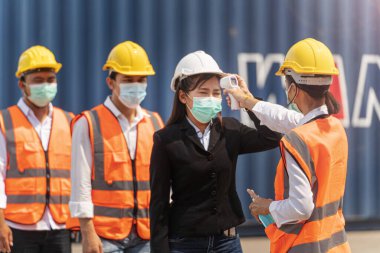 Yüz maskesi ve güvenlik elbisesi takan fabrika kadın çalışanı, temassız kızılötesi termometreyle kuyrukta bekleyen işçi sıcaklığını ölçüyor. kargo konteynırının arkaplanı.