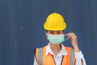 Asyalı kadın işçi mavi duvarda duruyor. İşçi kadın işçi koruyucu maske ve güvenlik kaskı takıyor ve turuncu yansıtıcı güvenlik elbisesi giyiyor. 