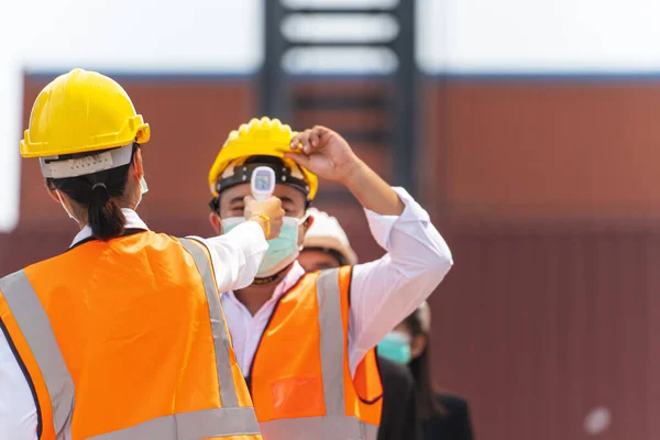 Factory woman worker in a face medical mask and safety dress used measures temperature at worker people standing on queue with a non-contact infrared thermometer. background of cargo container.