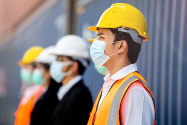 Grup işçileri koruma maskeleri ve güvenlik kasklarıdır. Arka planda kargo deposu vardır.