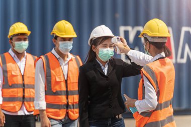 Yüz maskesi ve güvenlik elbisesi takan fabrika kadın çalışanı, temassız kızılötesi termometreyle kuyrukta bekleyen işçi sıcaklığını ölçüyor. kargo konteynırının arkaplanı.