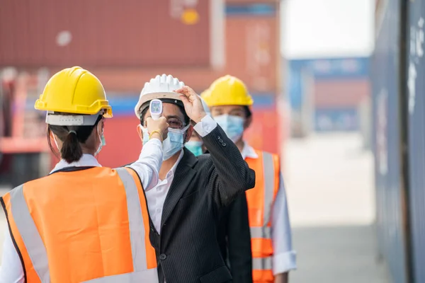 Factory woman worker in a face medical mask and safety dress used measures temperature at worker people standing on queue with a non-contact infrared thermometer. background of cargo container.