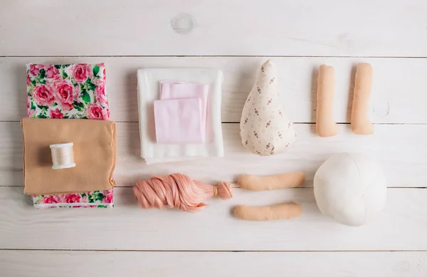 Textile doll in the sewing process with accessories and materials: fabric, thread, needle on white wooden background. Top view. Flat lay.