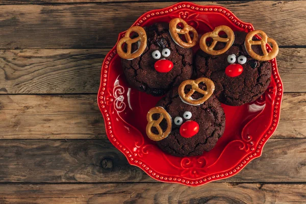 Reindeer cookies with candy red nose on a red plate. Christmas concept. Chocolate round cookies with a Christmas deer face with a red nose on a rustic wooden background. Top view