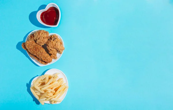 Plates in the shape of a heart with snacks on a blue background. Nuggets, wings, strips and fries with ketchup. Sunlight, harsh shadows. Top view with copy space