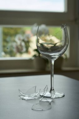 Kırık şarap kadehi ve cam kırıkları. Yüzeyde hasar görmüş cam parçaları var. Pencere manzaralı kapalı sahne.