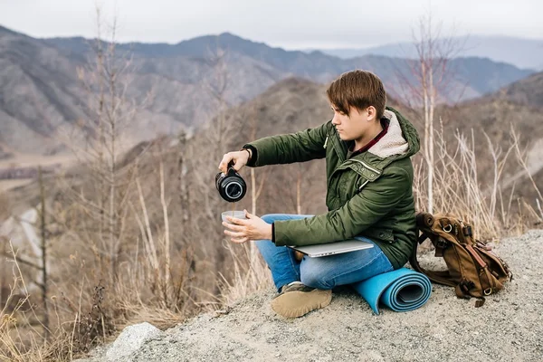 Man sitting on a background of mountains with a laptop pours tea from a thermos — Stock Photo, Image