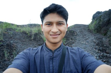 Siyah tişörtlü genç bir adamın portresi ve arka planda siyah kot pantolon ve mavi gökyüzü.