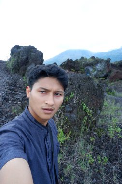 Siyah ceketli ve siyah elbiseli genç adam tepede dikiliyor ve uzaklığa bakıyor.