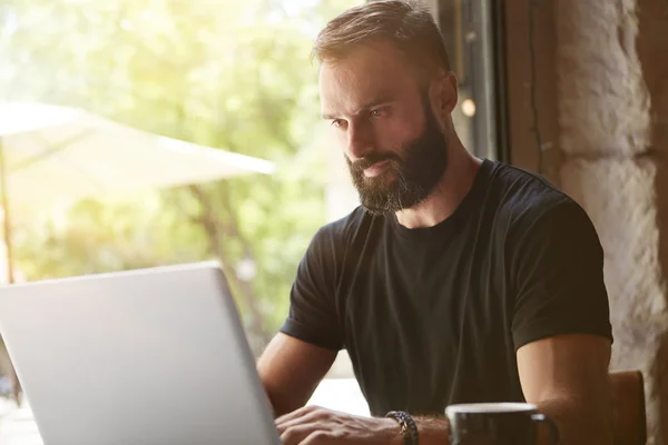 Koncentrerad Bearded man klädd i svart tshirt arbetar laptop träbord Urban Cafe. Young Manager arbete anteckningsbok modern inredning Loft Place. Coworking process Business Startup. suddig bakgrund. — Stockfoto