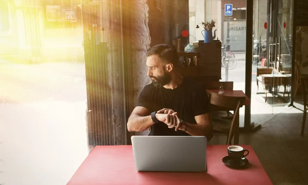 Young Bearded Businessman Wearing Black Tshirt Working Laptop Urban Cafe.Man Sitting Wood Table Cup Coffee Looking Through Window Touch Smartwatch.Coworking Process Business Startup.Blurred Background — Stockfoto
