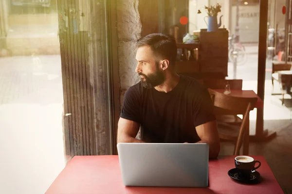 Ung skäggig affärsman bär svart tröja arbetar laptop Urban Cafe.Man sitter trä tabell Cup kaffe tittar genom fönstret.Coworking Process Business Startup.suddig bakgrund. solljus effekt. — Stockfoto