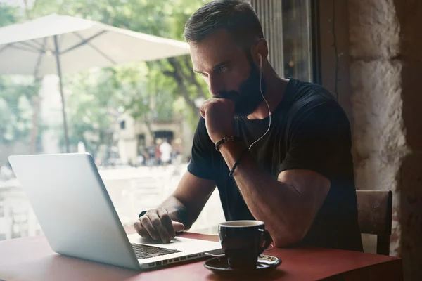 Koncentrerad ung skäggiga affärsman bär svart tshirt arbetar laptop Urban Cafe. man sittande träbord kopp kaffe tittar genom window. Coworking process Business Startup. suddig bakgrund — Stockfoto
